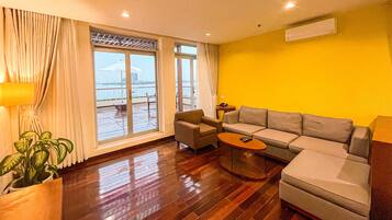 A living room with a sofa, armchair, and coffee table. There is a large window with curtains, a potted plant, and an air conditioning unit on the wall.