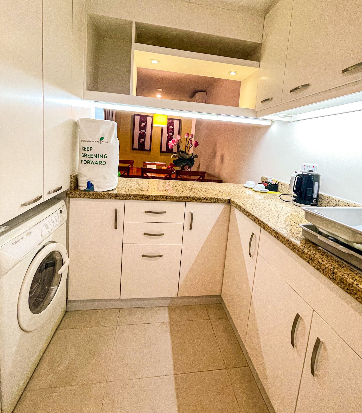 A compact kitchen with a washing machine, a countertop with a coffee maker, and a bag with a message.