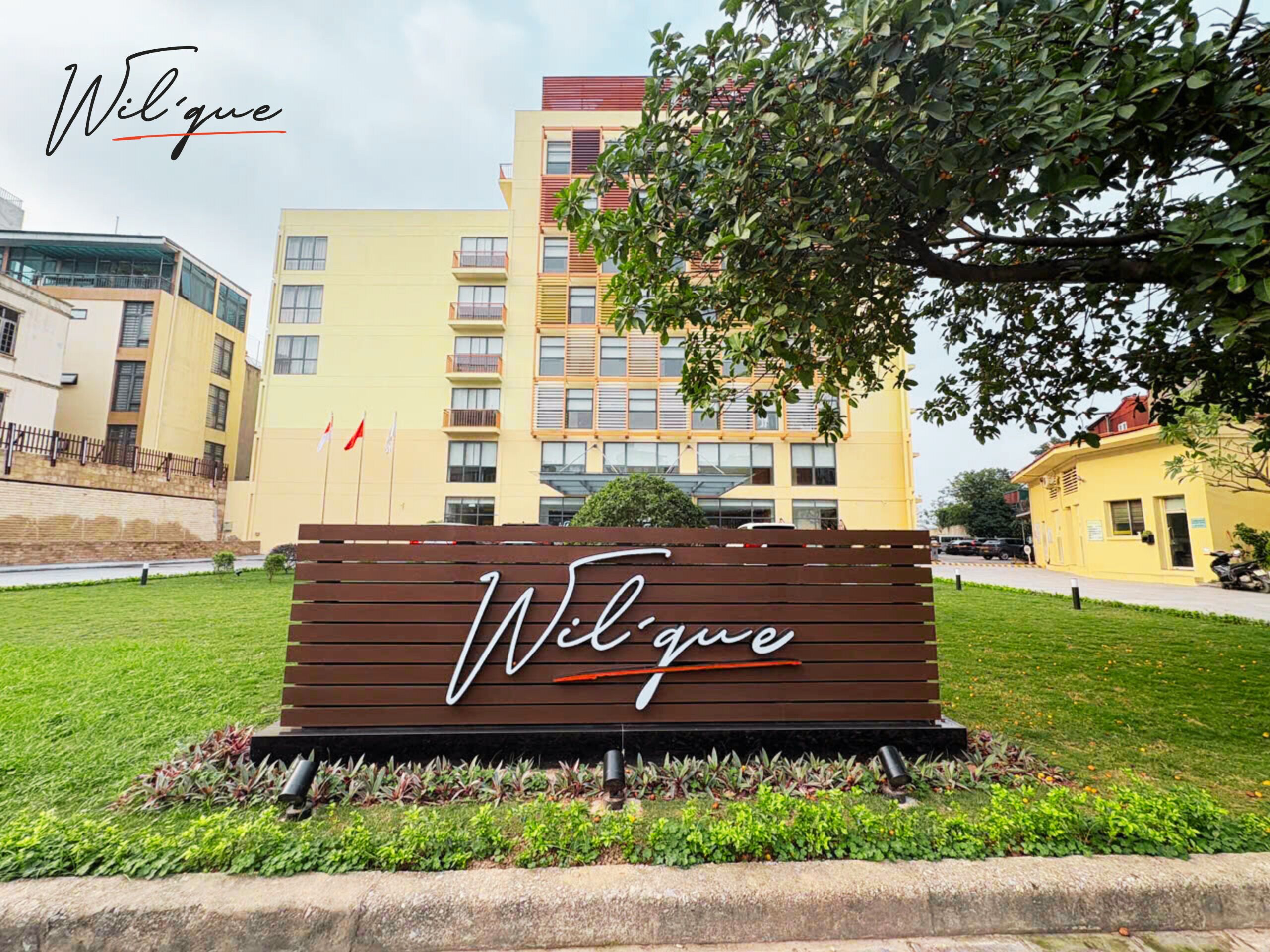 A sign with the name 'Wilque' in a modern font, surrounded by greenery and a building complex in the background.