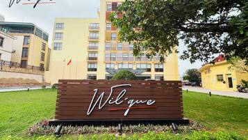 A sign with the name 'Wilque' in a modern font, surrounded by greenery and a building complex in the background.