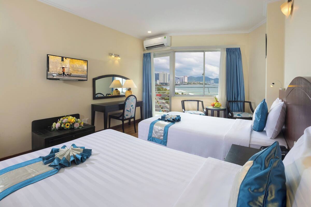 Ein Hotelzimmer mit zwei Betten, einem Schreibtisch, einem Stuhl, einem kleinen Tisch mit Früchten und einem großen Fenster mit Blick auf die Stadt.