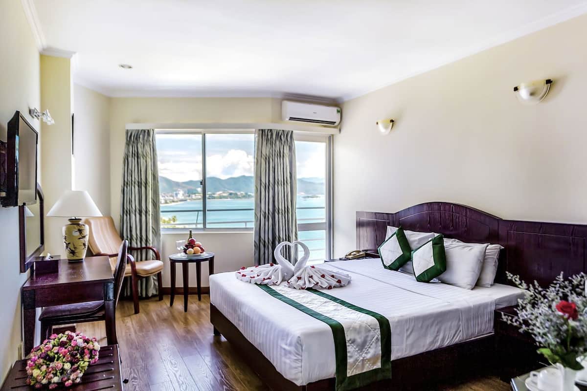 Quarto de hotel com cama, escrivaninha, cadeira, luminária, um vaso com flores e vista para o mar.