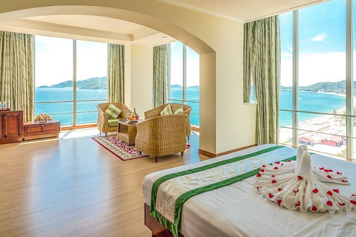 Quarto de hotel com uma cama grande, área de estar com cadeiras, penteadeira de madeira e vista para o mar.