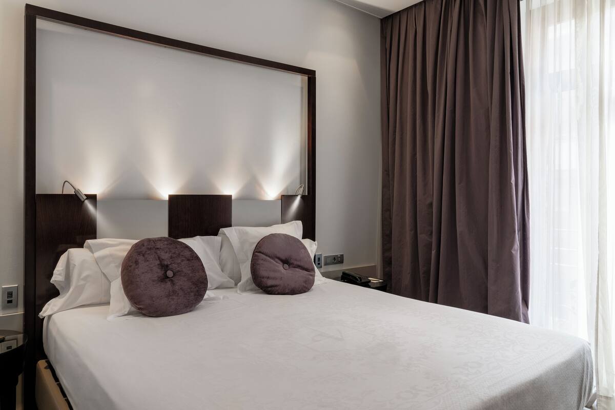 A neatly made bed with white linens and two large pillows, a wooden headboard with integrated lighting, a nightstand with a lamp, and a window with sheer curtains.