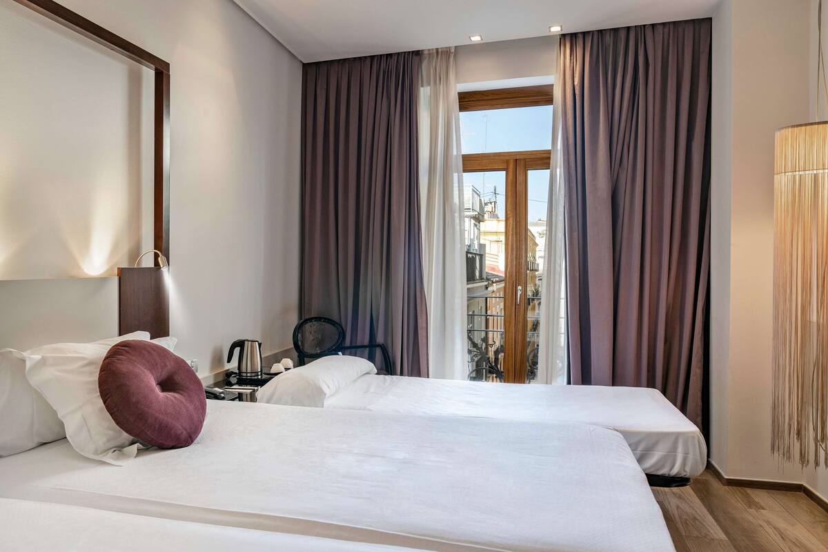 A hotel room with two beds, a large window with curtains, a view of buildings outside, and a lamp on the nightstand.