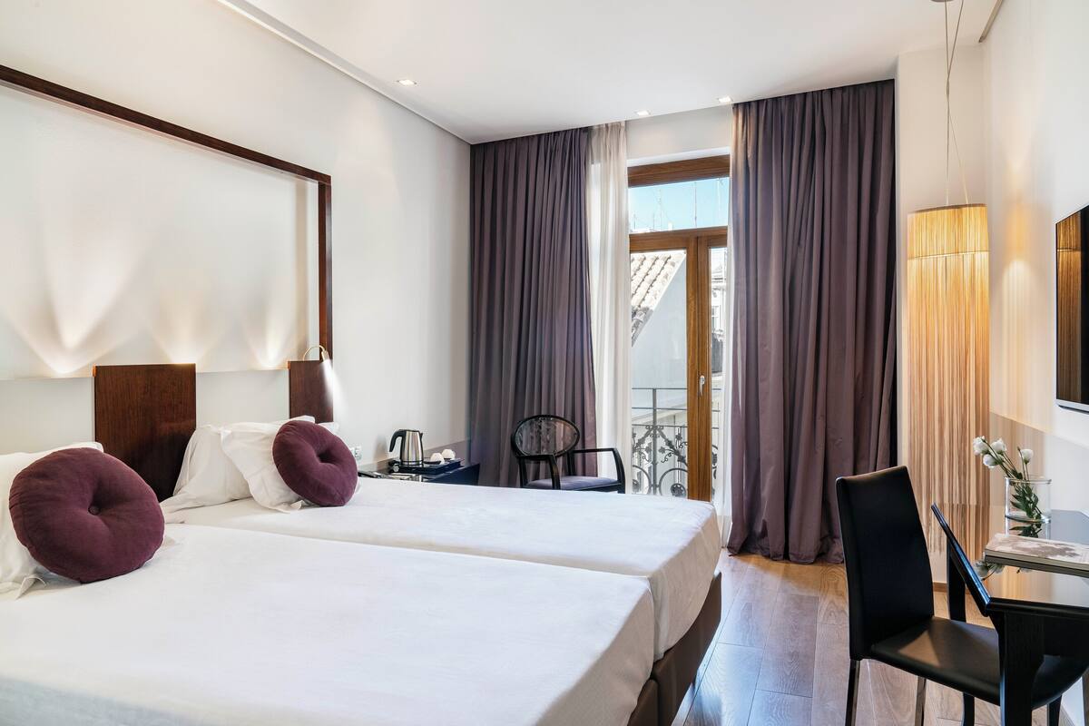 A hotel room with two beds, a desk, a chair, and a balcony with curtains.