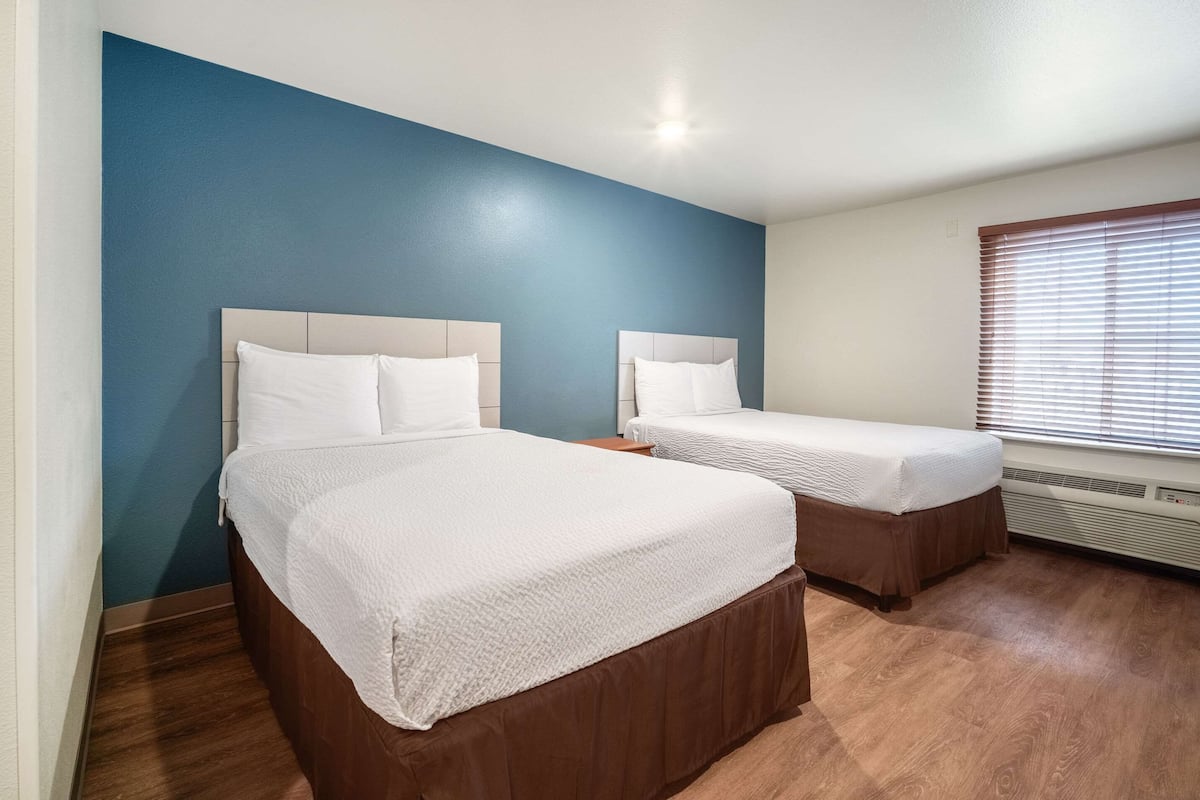 A hotel room with two beds, a blue accent wall, wooden flooring, and a window with blinds.