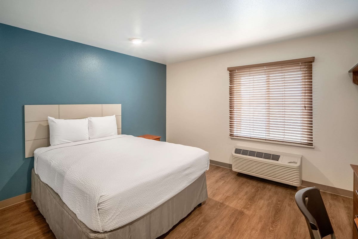 A bedroom with a bed, a nightstand, a window with blinds, and a wall-mounted heater.