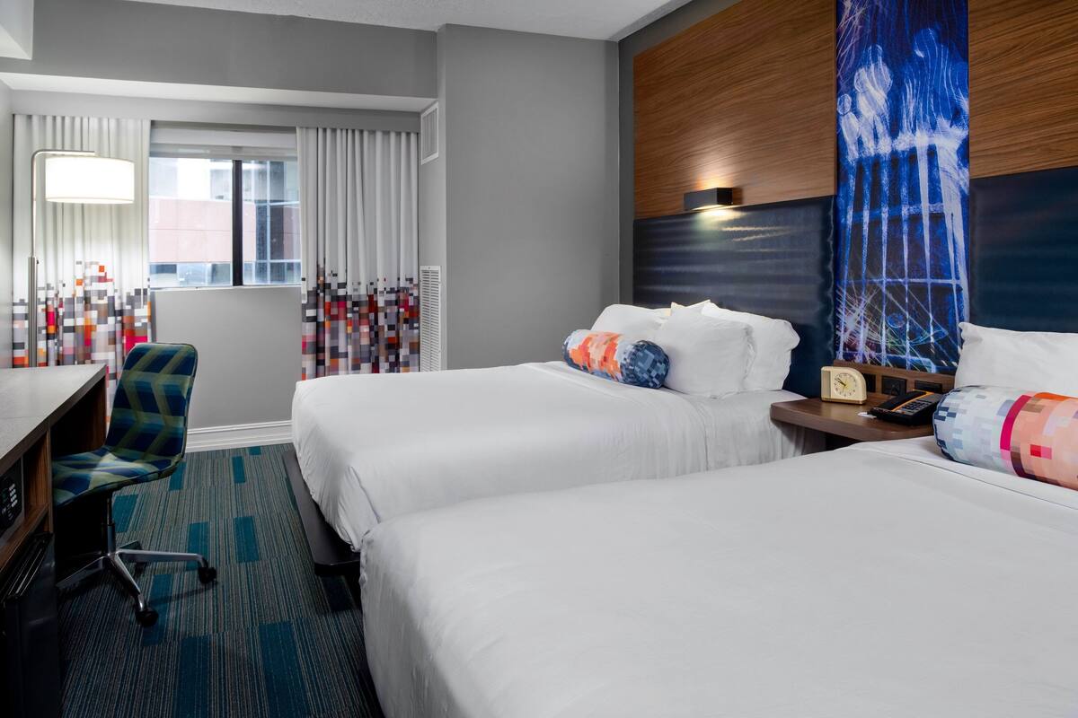 A hotel room with two beds, a desk, a chair, a large window with curtains, and a wall with a textured design.