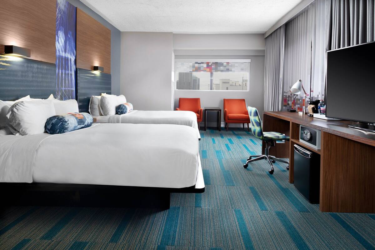 A hotel room with a large bed, two armchairs, a desk with a television, and a city view through the window.