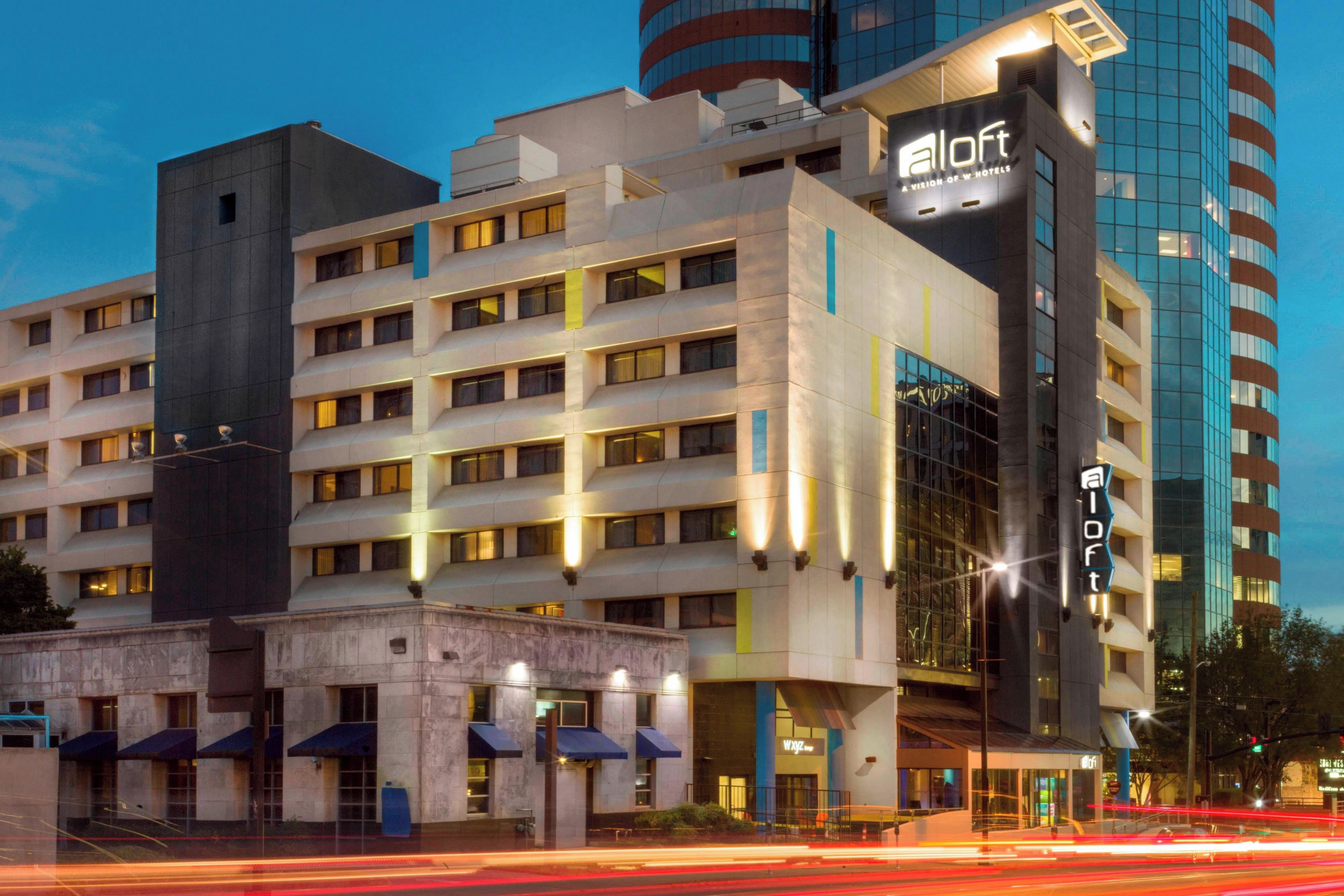 A modern hotel building with multiple floors, a prominent 'aloft' sign, and a glass facade reflecting the sky.