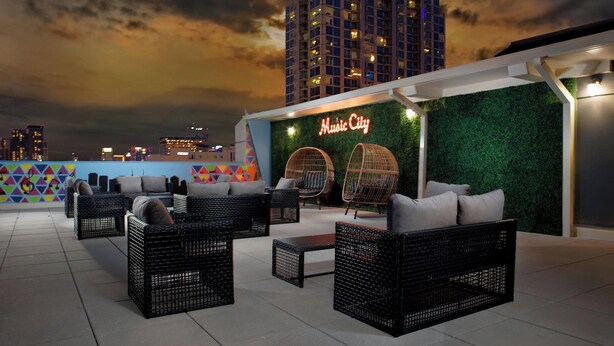 Un área de estar al aire libre con mobiliario de mimbre y un letrero de neón que dice "Music City" sobre un fondo verde.