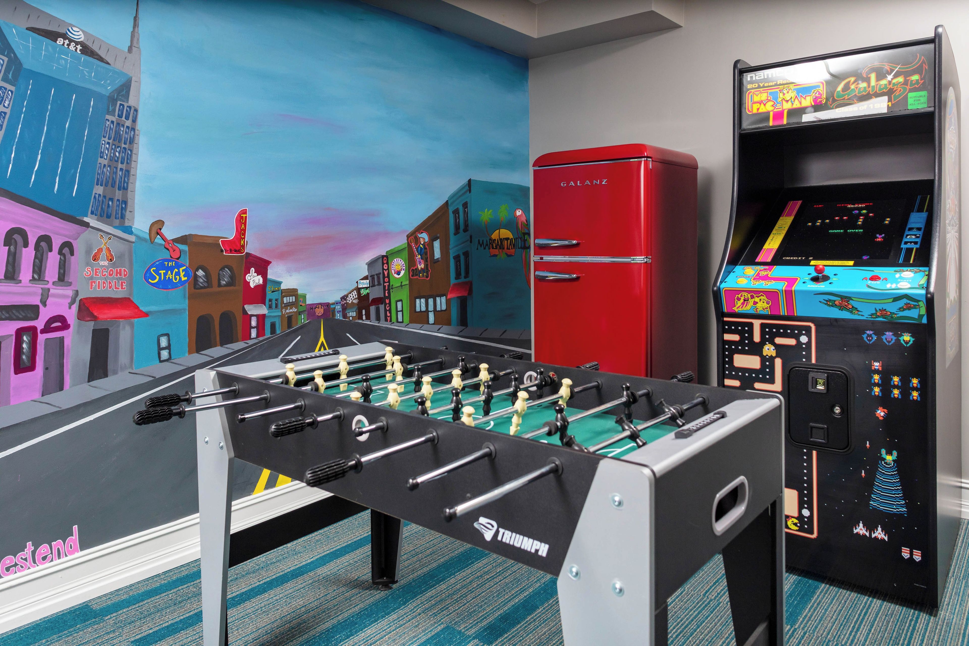 Une pièce avec un baby-foot, un réfrigérateur rouge et une console de jeux.
