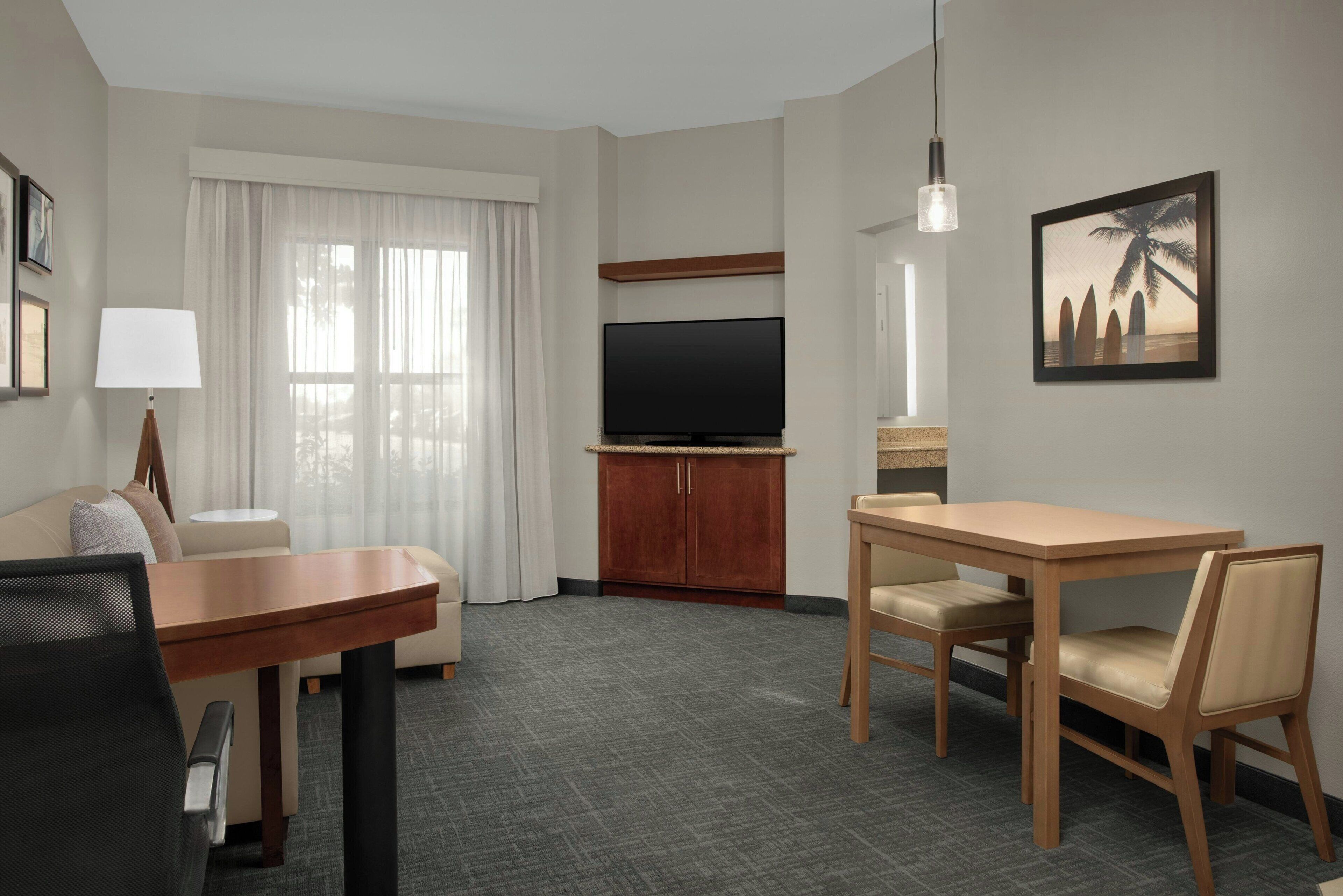 A hotel room with a flat-screen TV, a dining table with chairs, a sofa, a lamp, and a painting on the wall.