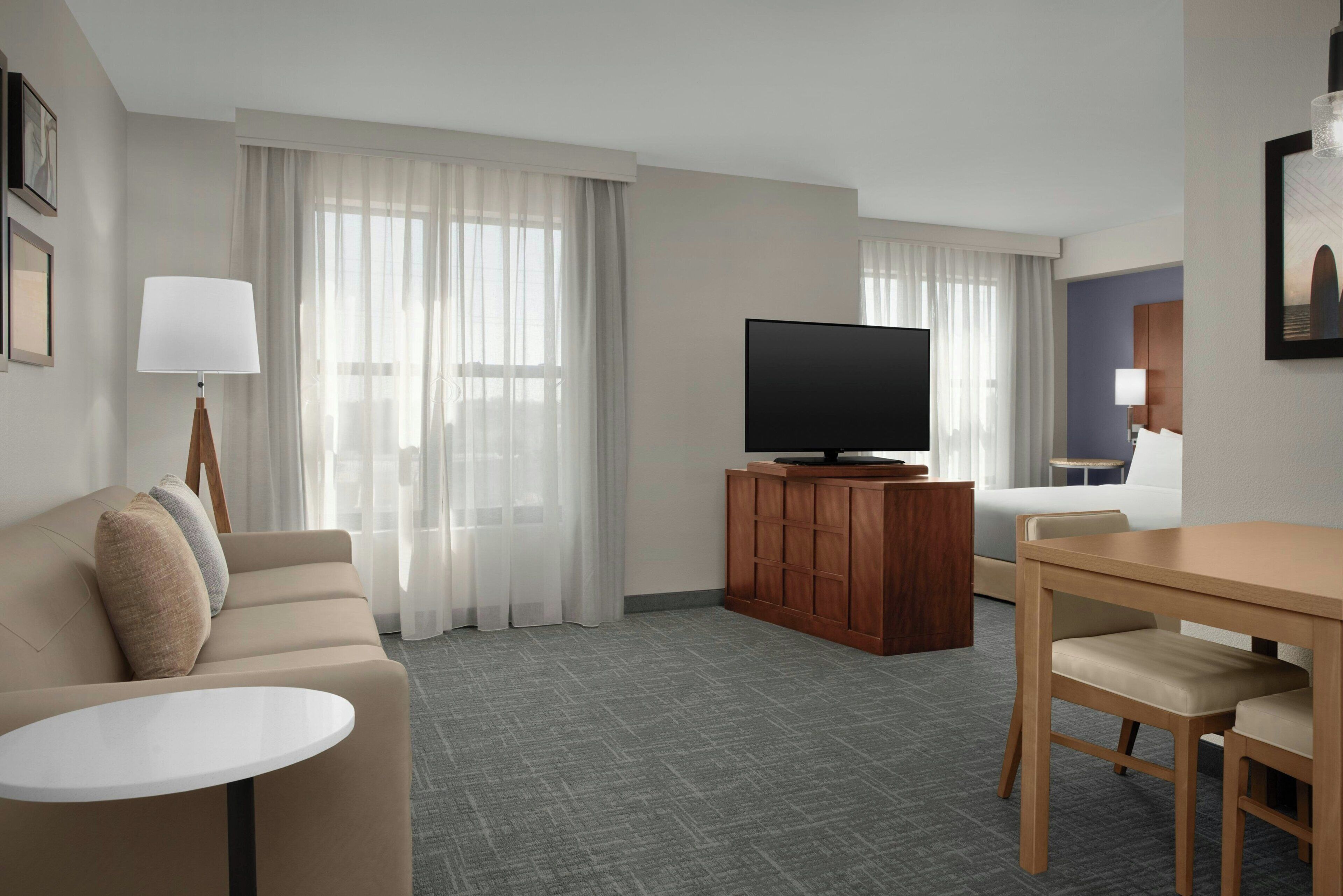 A hotel room with a sofa, a TV on a wooden stand, a dining table with chairs, and a large window with sheer curtains.