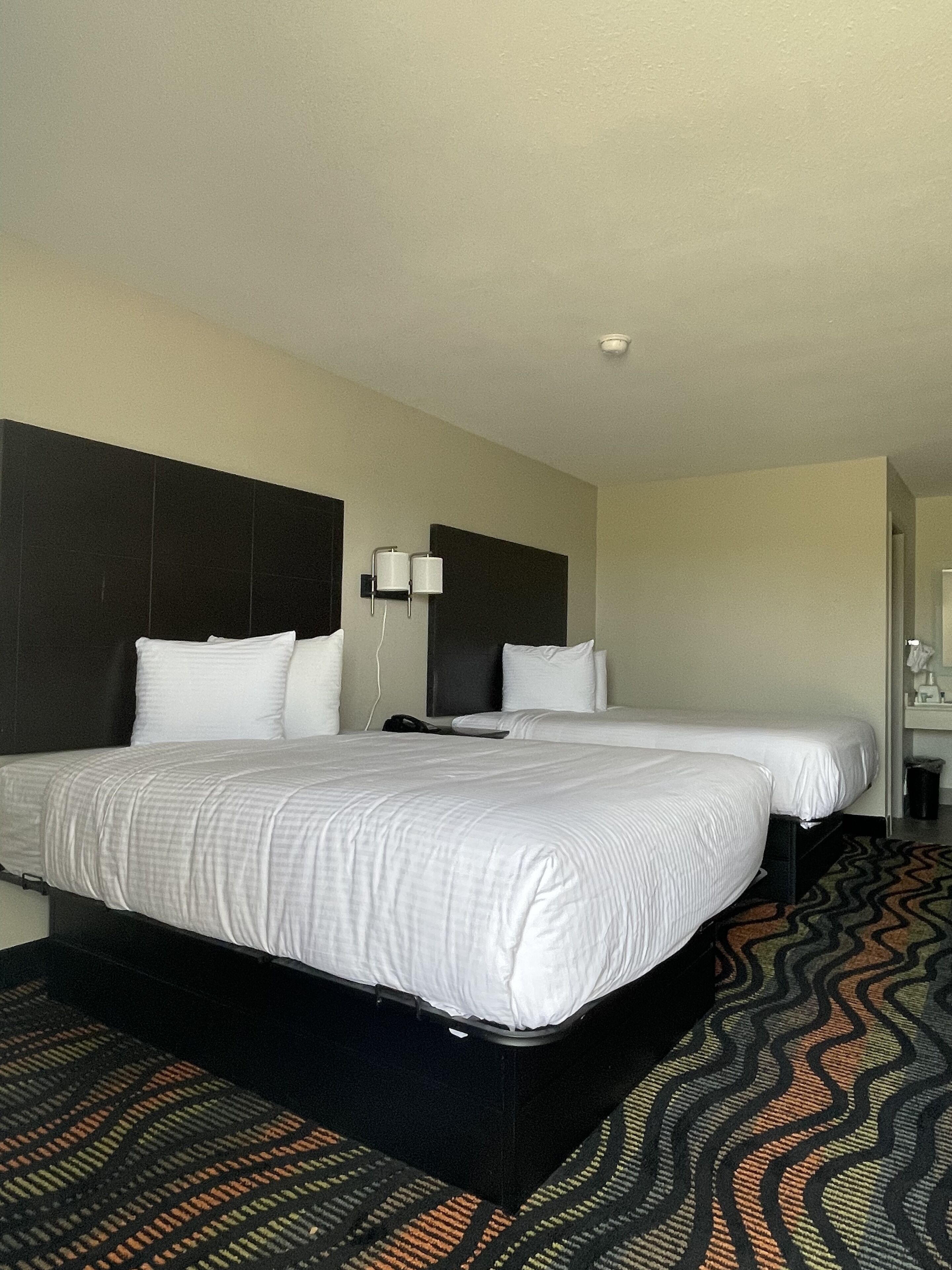 A hotel room with a large bed, a smaller bed, a nightstand, a lamp, and a bathroom visible in the background.