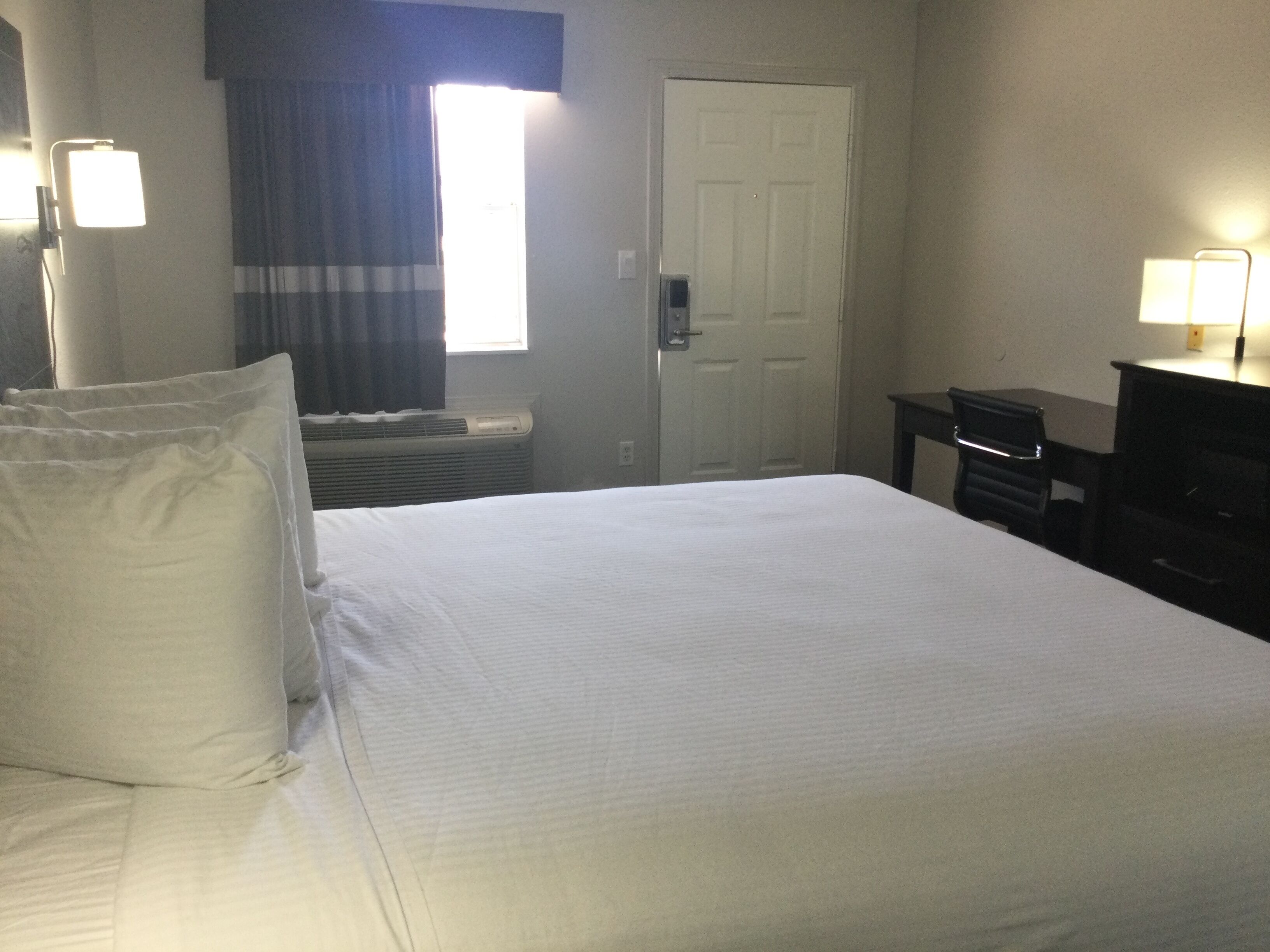 Une chambre d’hôtel avec un lit, un bureau, une chaise, une porte et une fenêtre avec des rideaux.