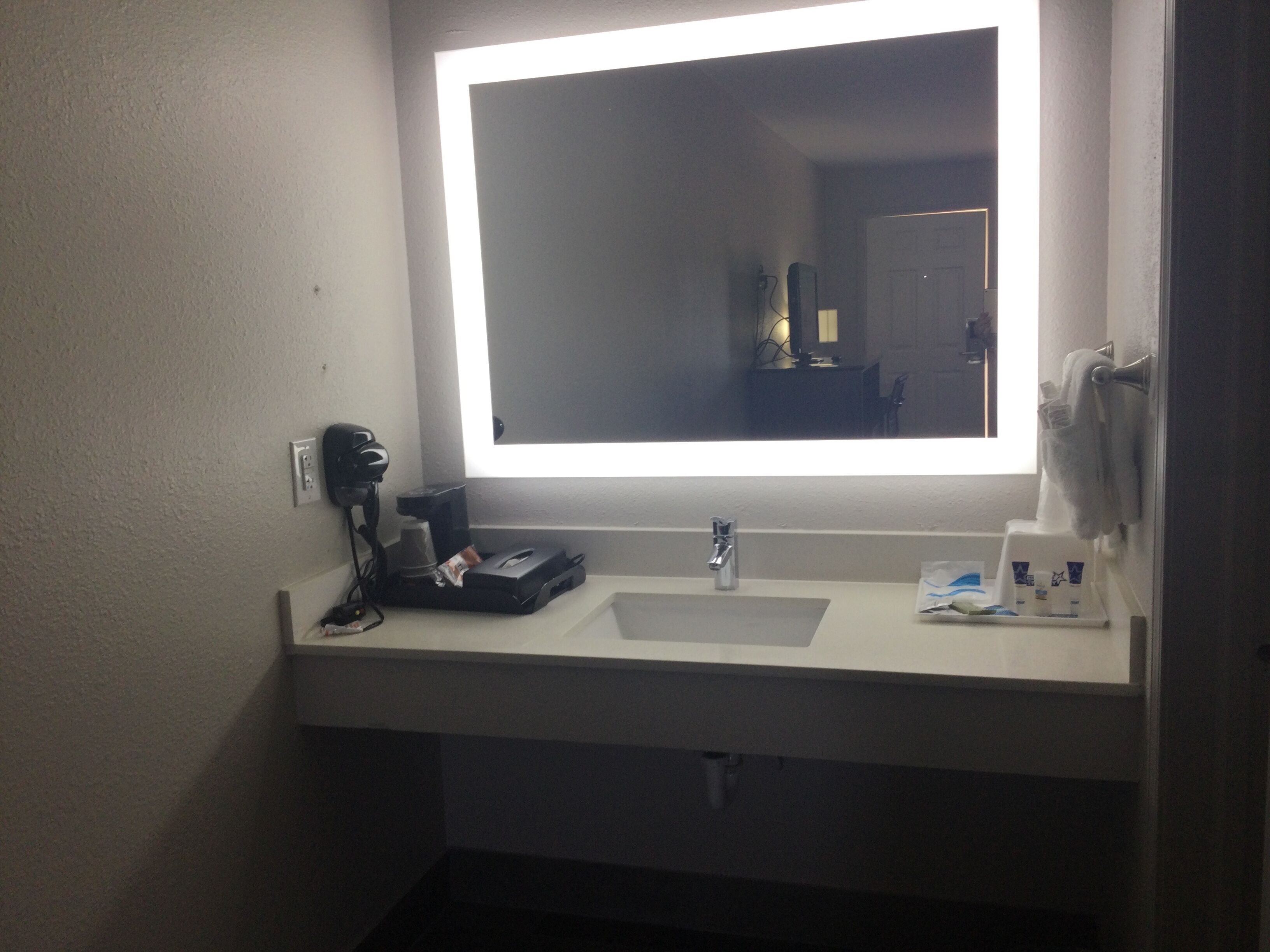 A bathroom with a white sink, a mirror with a bright light, a hairdryer, and a towel rack.