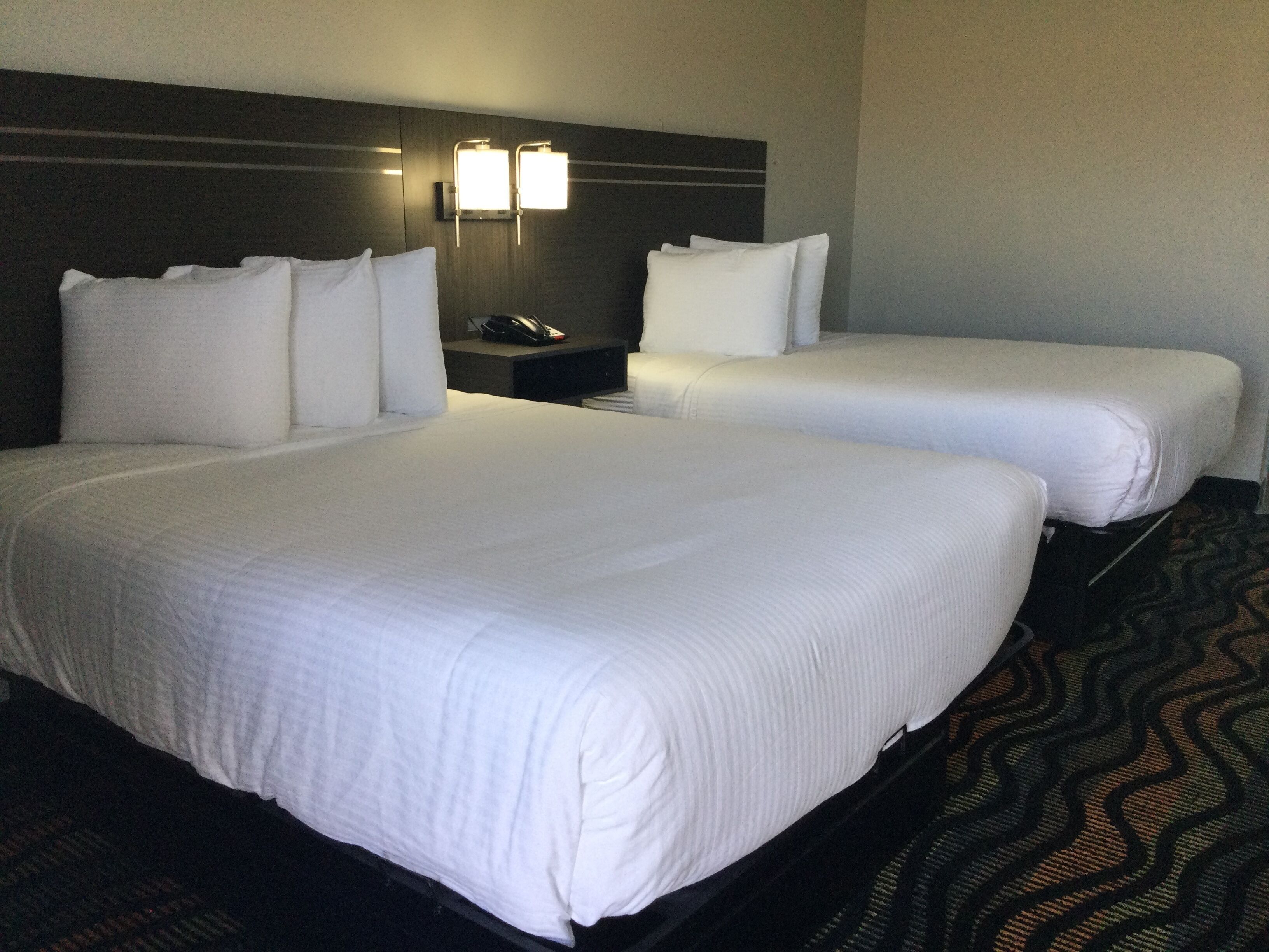 A hotel room with two beds, a nightstand, and a wall-mounted lamp.