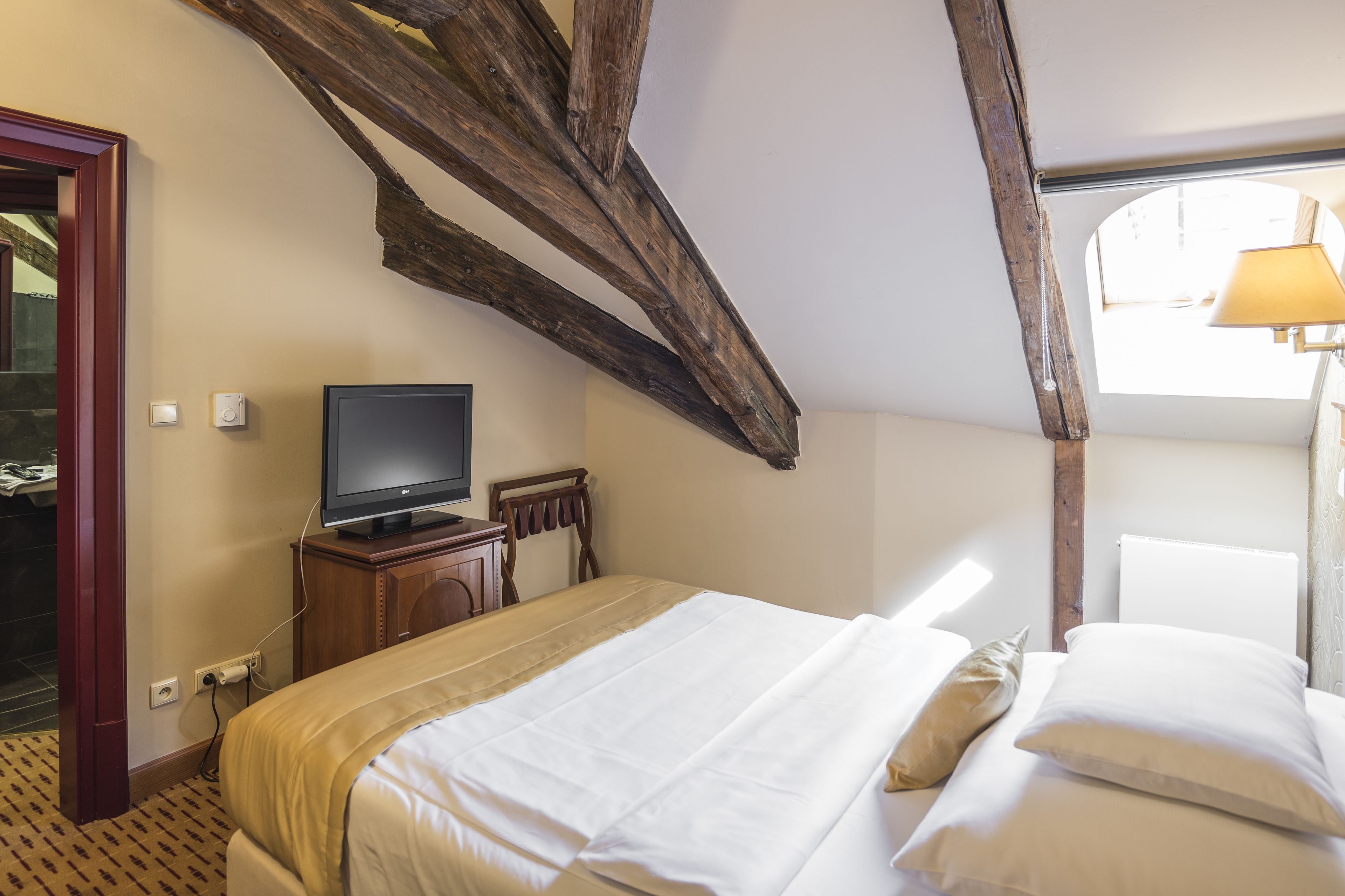 A bedroom with a wooden ceiling, a bed with white bedding, a television on a wooden cabinet, and a window with natural light.