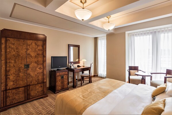 A hotel room with a large bed, a desk with a television, a wardrobe, and a seating area with two chairs. - Majestic Plaza Hotel Prague (Prague)