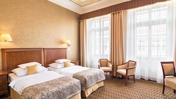 Habitación de hotel con dos camas, cabecero de madera, un ventanal con cortinas y alfombra estampada.
