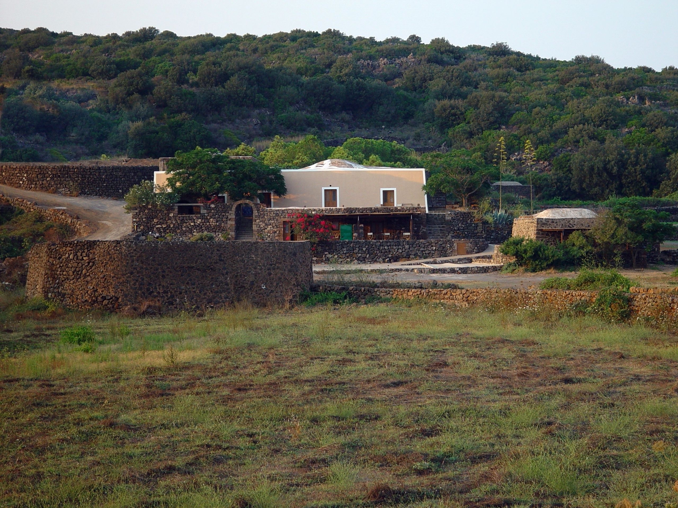 A stone house with a flat roof, surrounded by a stone wall and a dirt path.
