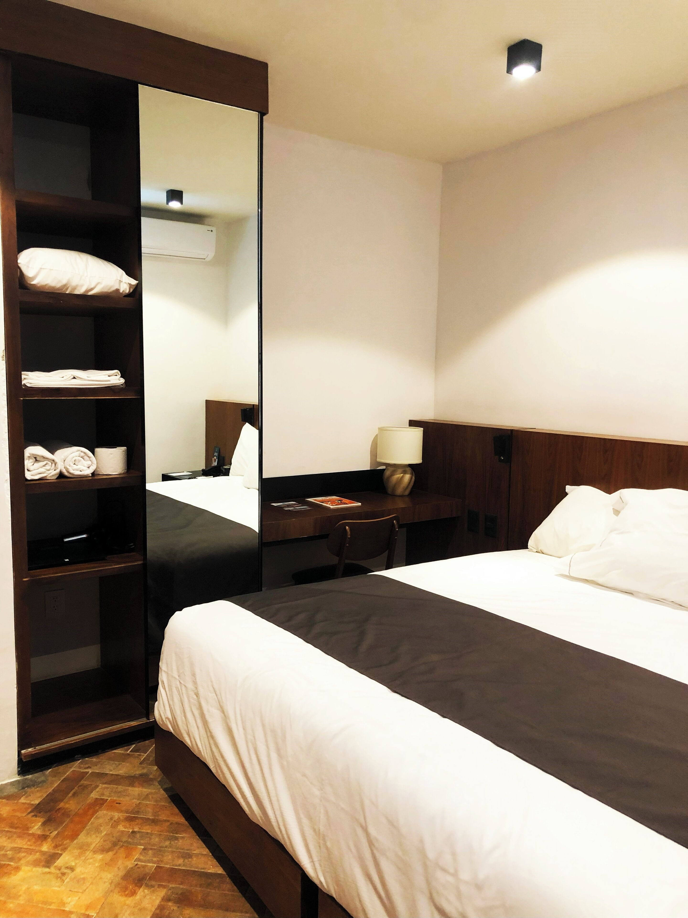 A hotel room with a bed, a desk, a chair, a mirror, and a wardrobe with folded towels.