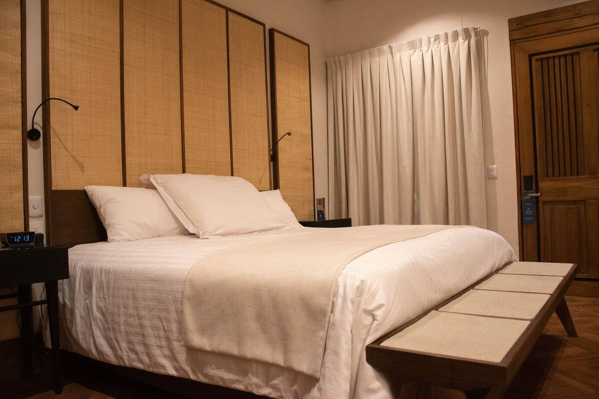 Un lit bien fait, avec du linge de lit blanc, une table de chevet avec une horloge et un banc en bois au pied du lit.