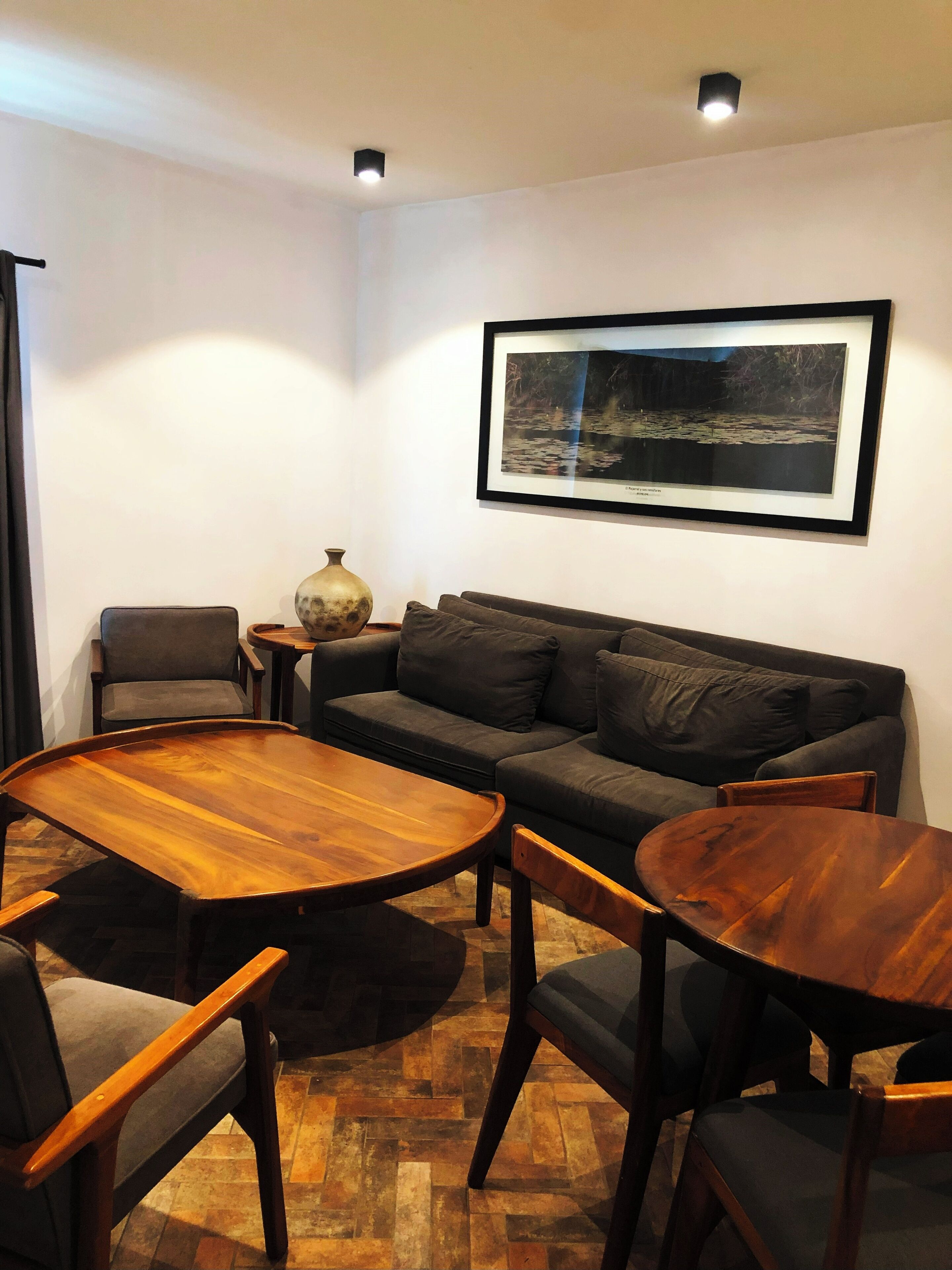 Uma sala de estar moderna com um sofá cinza escuro, duas mesas de centro de madeira e cadeiras combinando. Uma fotografia emoldurada está pendurada na parede acima do sofá.