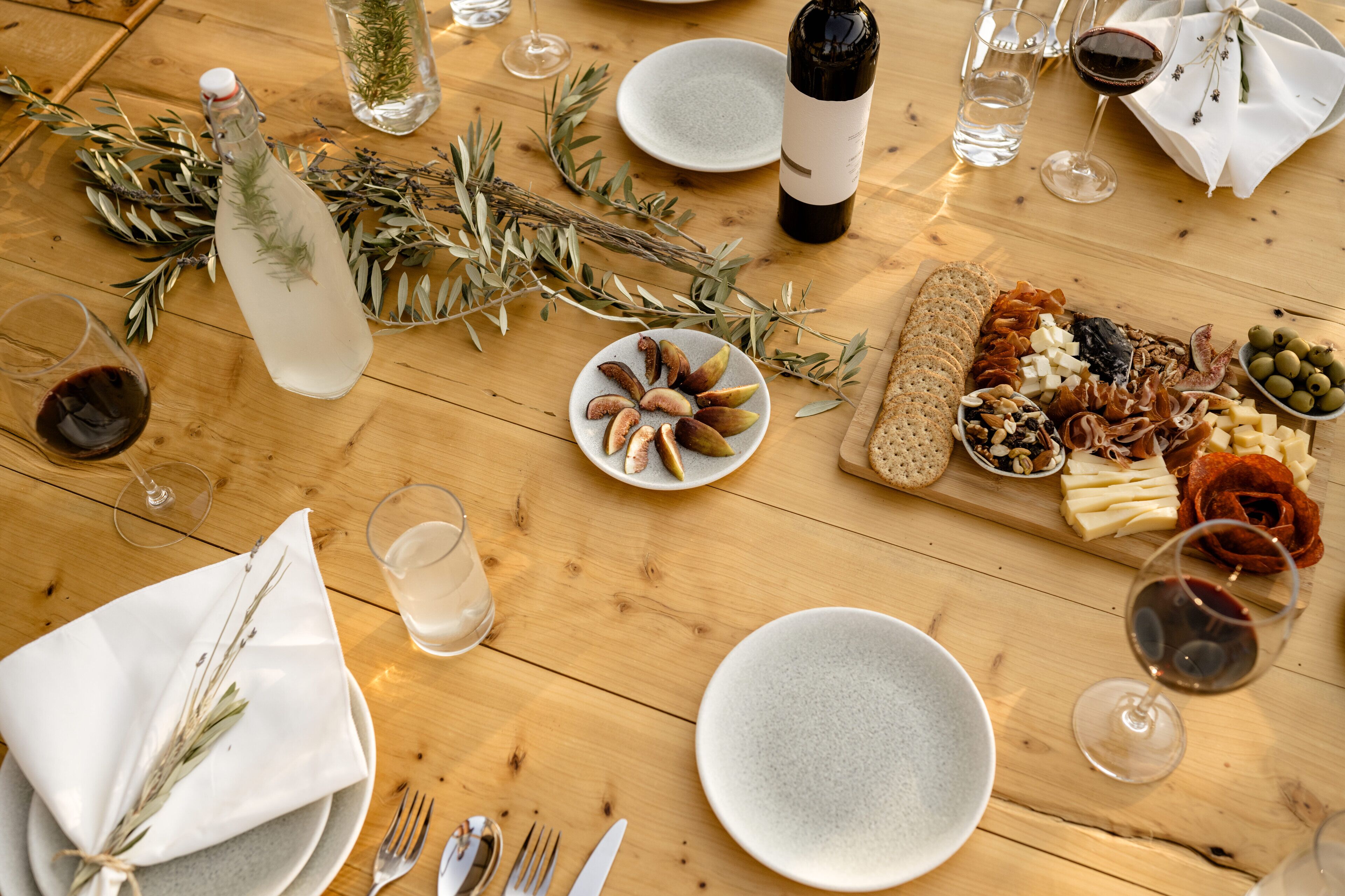 A wooden table set for a meal with plates, cutlery, a bottle of water, a bottle of wine, and a charcuterie board.