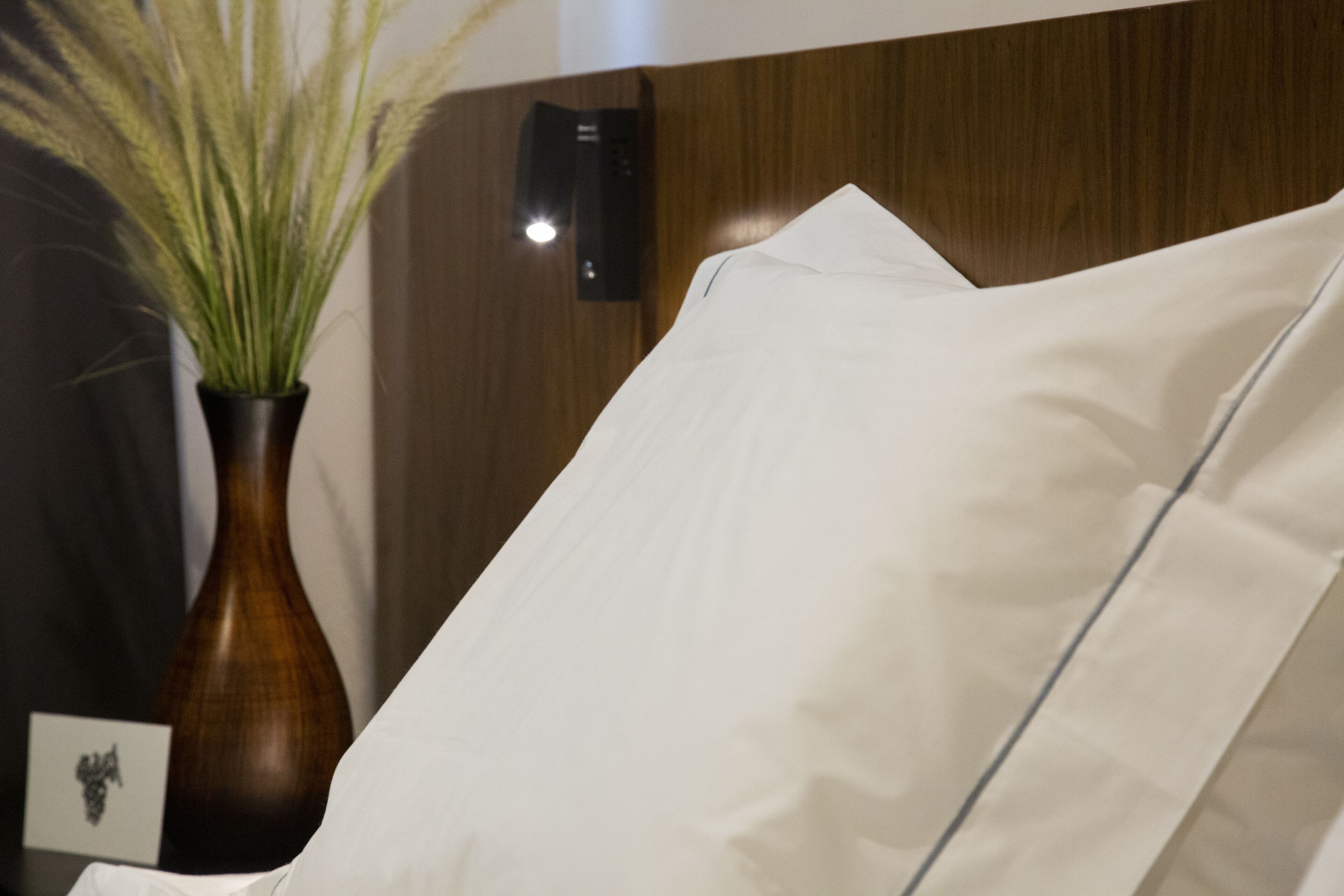 Una cama con sábanas blancas, un cabecero de madera, un florero con plantas verdes y un dispositivo montado en la pared.