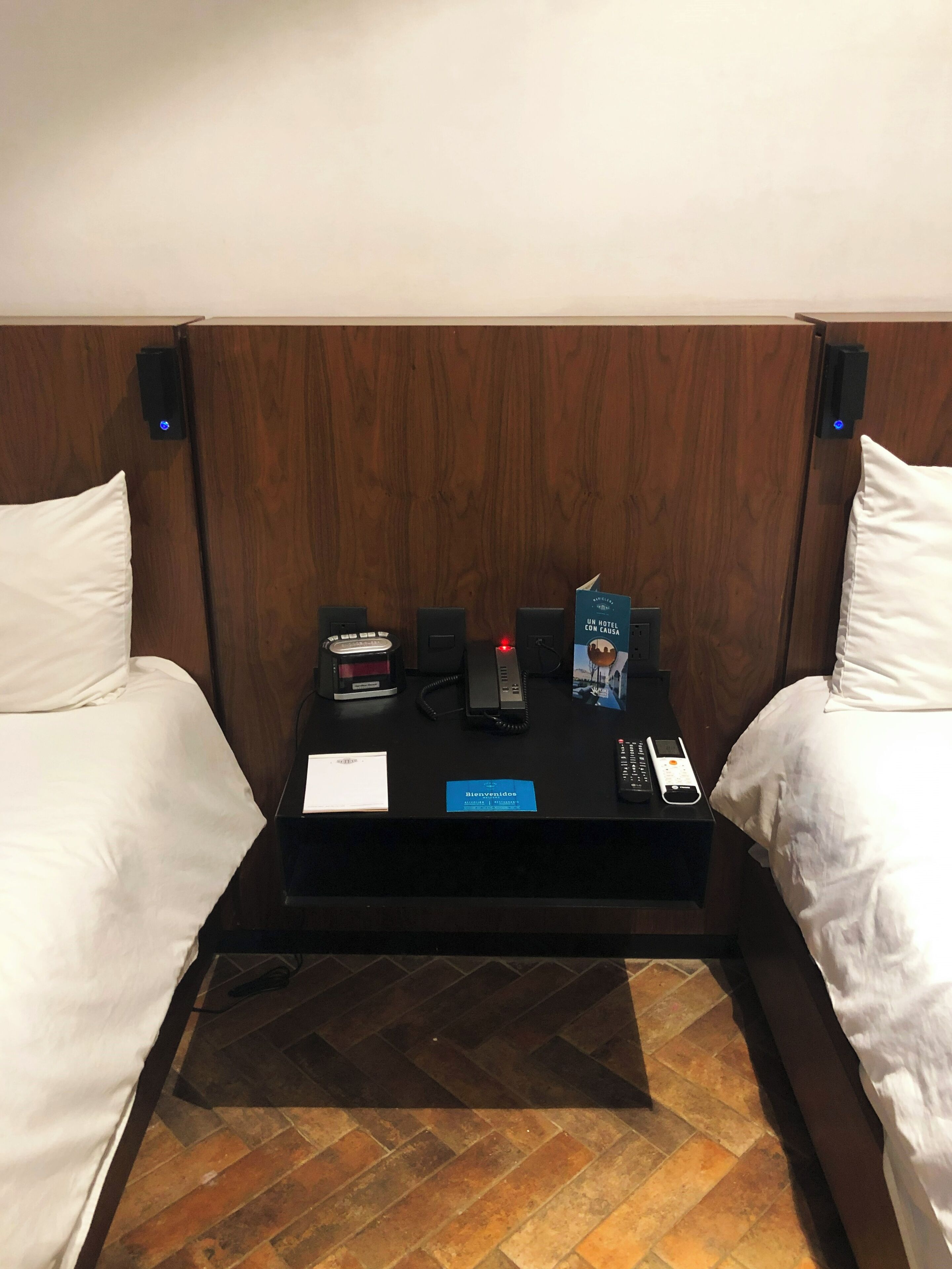 A hotel room with a wooden headboard, two white beds, a small wooden table with a welcome card, a phone, a remote control, and a small box.