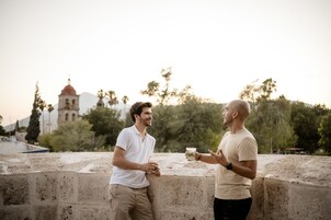 Dos hombres conversando en un saliente de piedra con una iglesia al fondo.