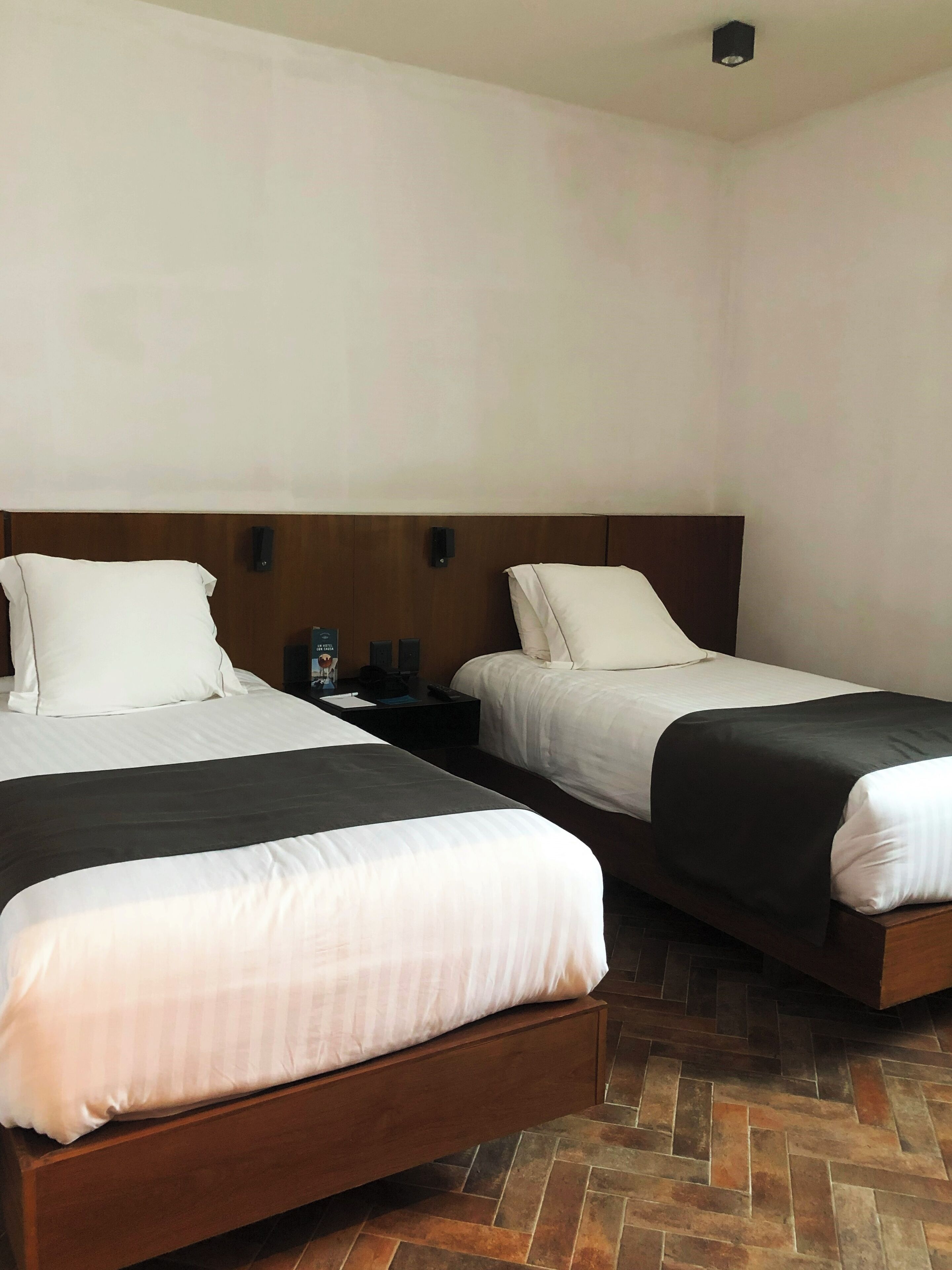 A hotel room with two single beds, wooden headboards, a nightstand with a lamp, and a herringbone-patterned floor.