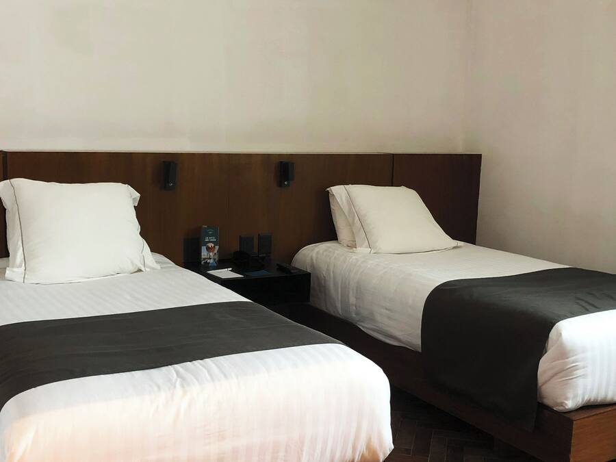A hotel room with two single beds, wooden headboards, a nightstand with a lamp, and a herringbone-patterned floor.