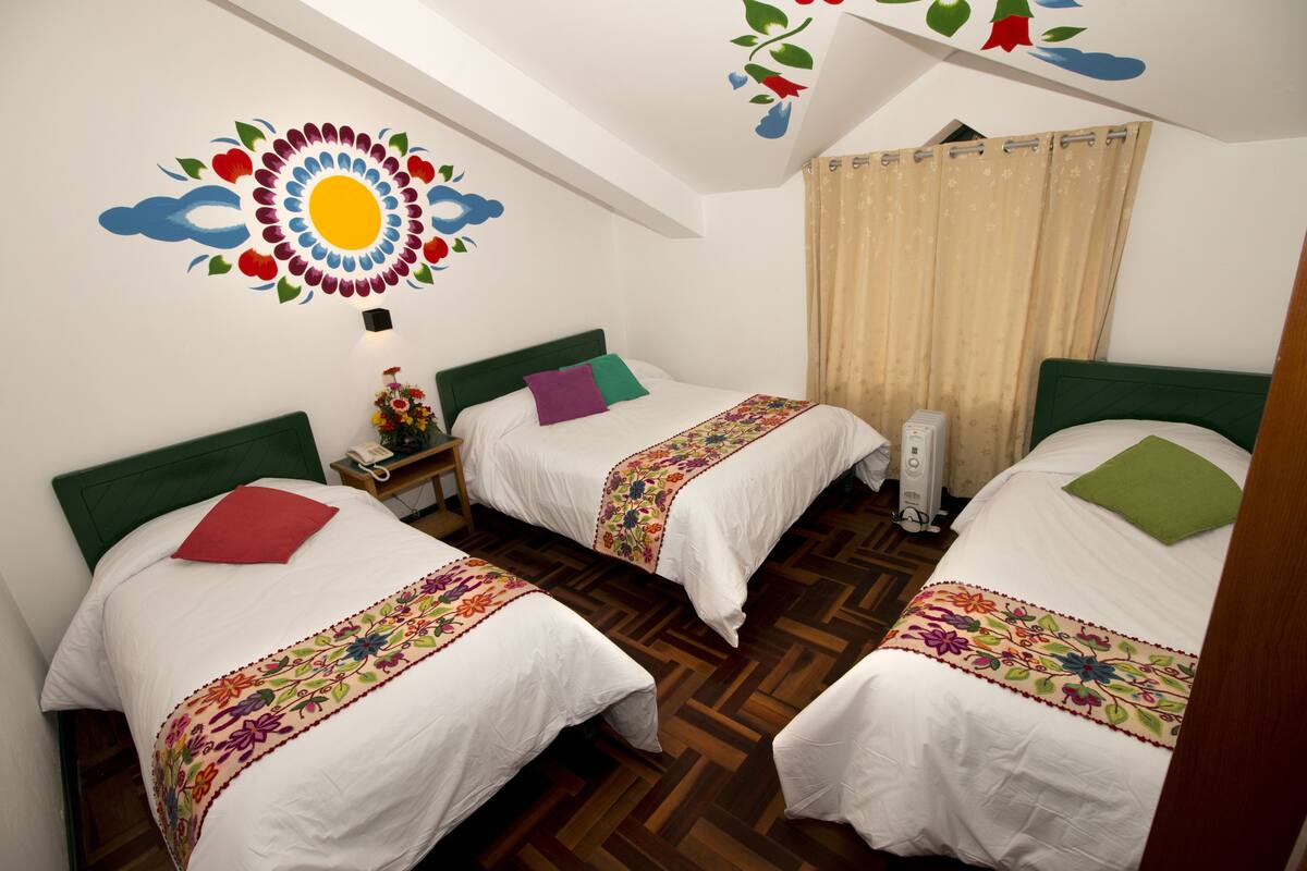A room with two beds, each with a green headboard, white bedding, and colorful floral bedspreads. There is a ceiling with a sun and cloud mural, and a window with beige curtains.