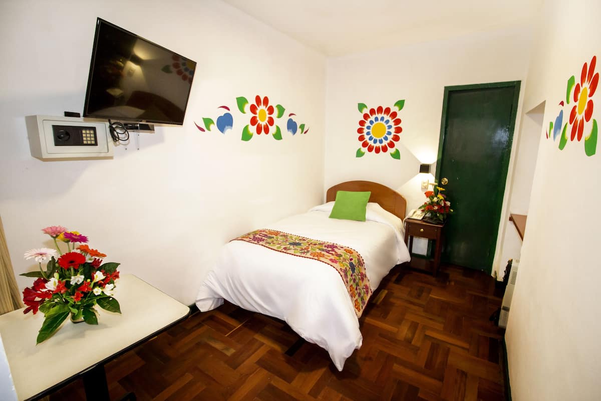 A bedroom with a bed, a TV mounted on the wall, a desk with a vase of flowers, and a door with a green frame.