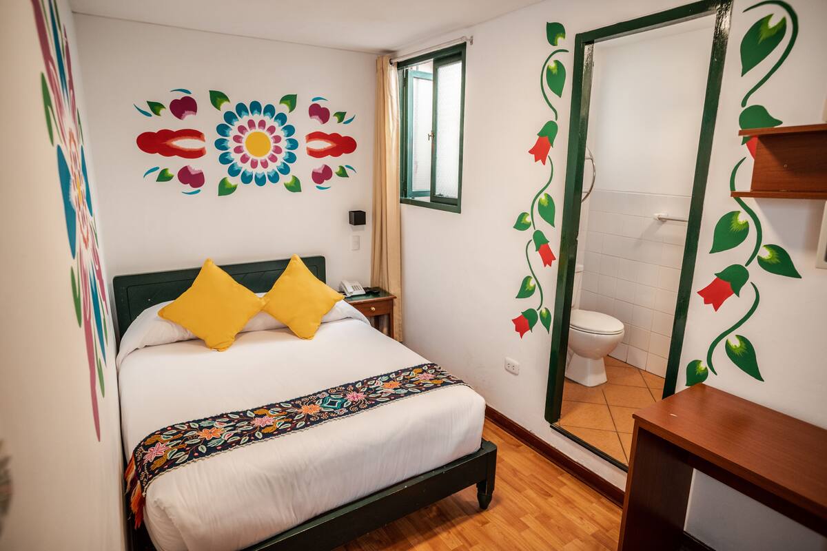 A neatly made bed with a floral-patterned bedspread and two pillows, a wooden nightstand with a telephone, and a bathroom with a white toilet and a green-framed mirror.