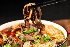 A bowl of noodle soup with meat and vegetables, being lifted with chopsticks.