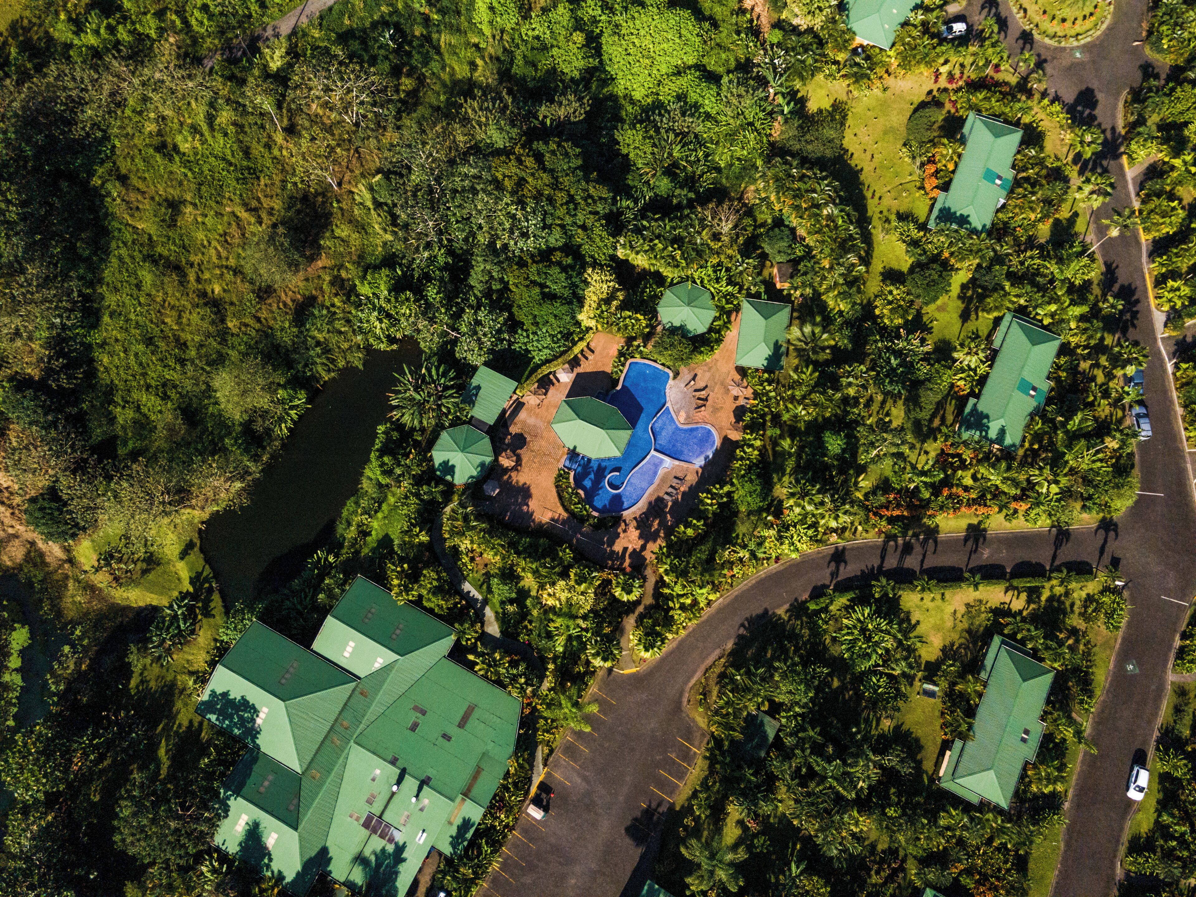 An aerial view of a resort with multiple green-roofed buildings, a central swimming pool, and surrounding lush greenery.