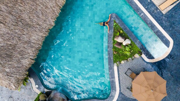 An aerial view of a swimming pool with a person swimming, surrounded by a thatched roof, lounge chairs, and an umbrella.