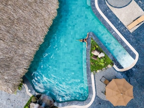 An aerial view of a swimming pool with a person swimming, surrounded by a thatched roof, lounge chairs, and an umbrella. - Arenal Manoa & Hot Springs Resort (La Fortuna)
