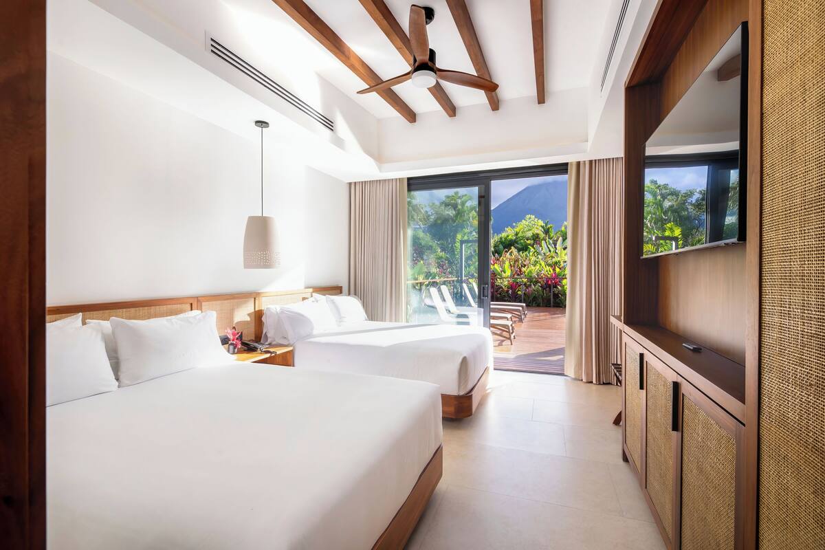 A hotel room with two beds, a ceiling fan, a large window with a view of greenery, and a TV on a wooden cabinet.