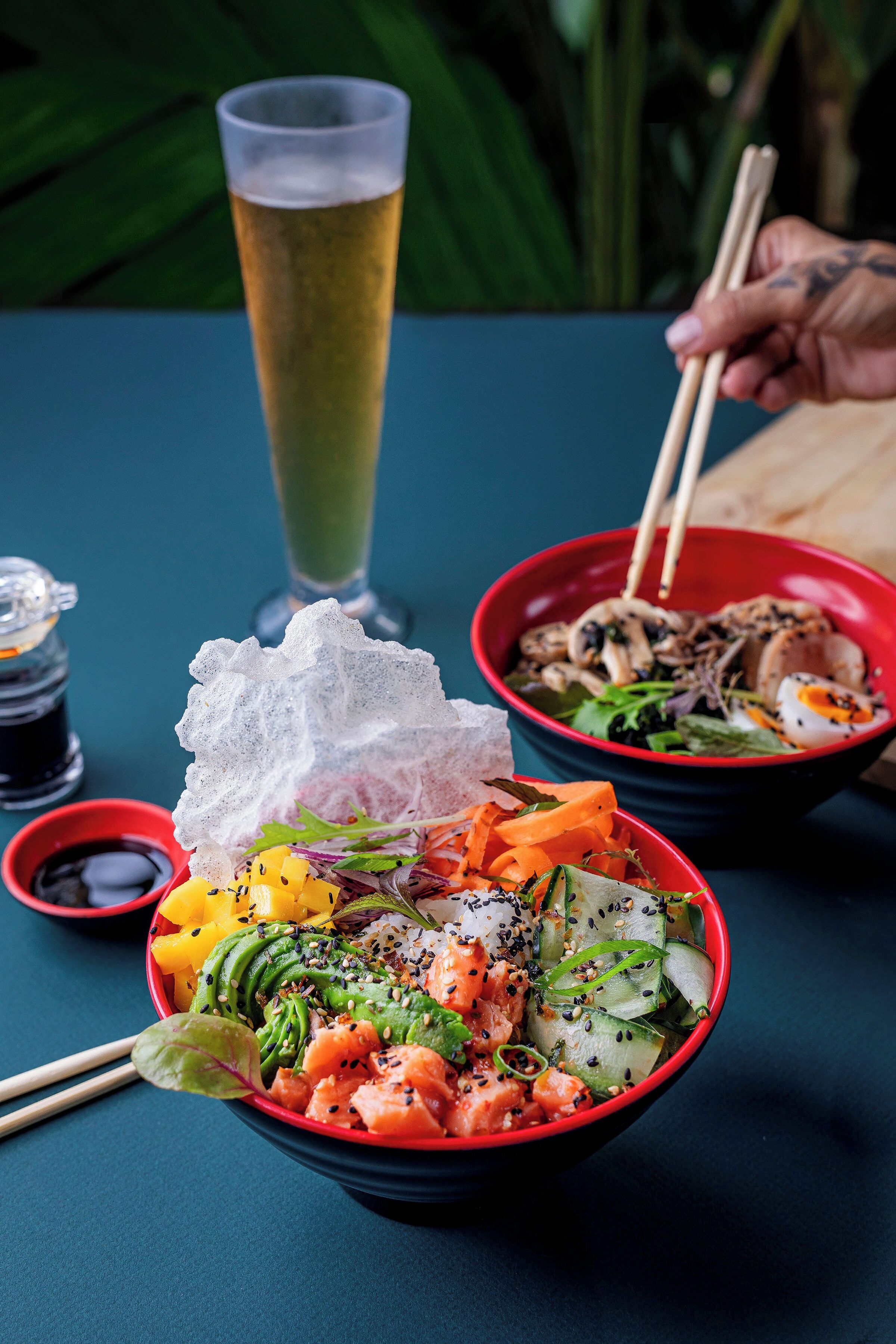 A table with a bowl of sushi, a glass of beer, and a red bowl with a salad.