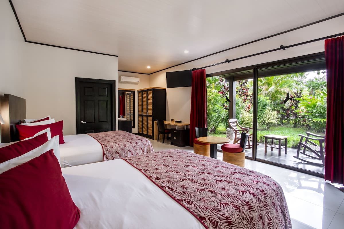 A hotel room with two beds, a dining area, and a view of a lush garden.