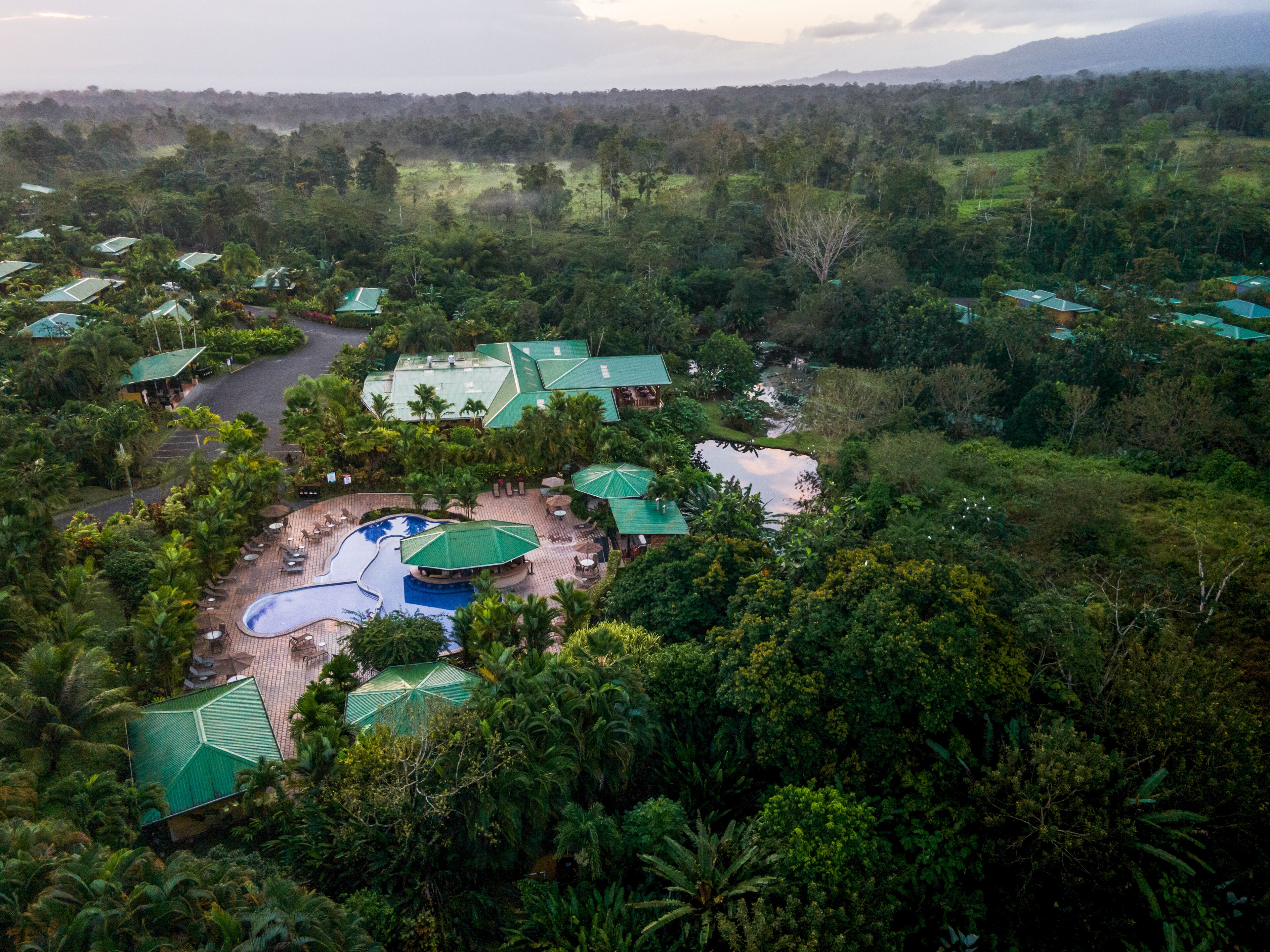 An aerial view of a resort with multiple green-roofed buildings, a swimming pool, and lush surrounding vegetation.