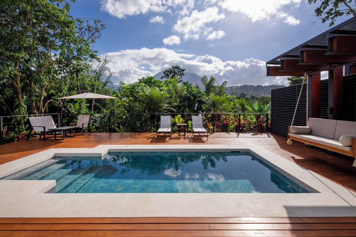 A swimming pool with lounge chairs and a wooden deck, surrounded by lush greenery and a mountain in the background.