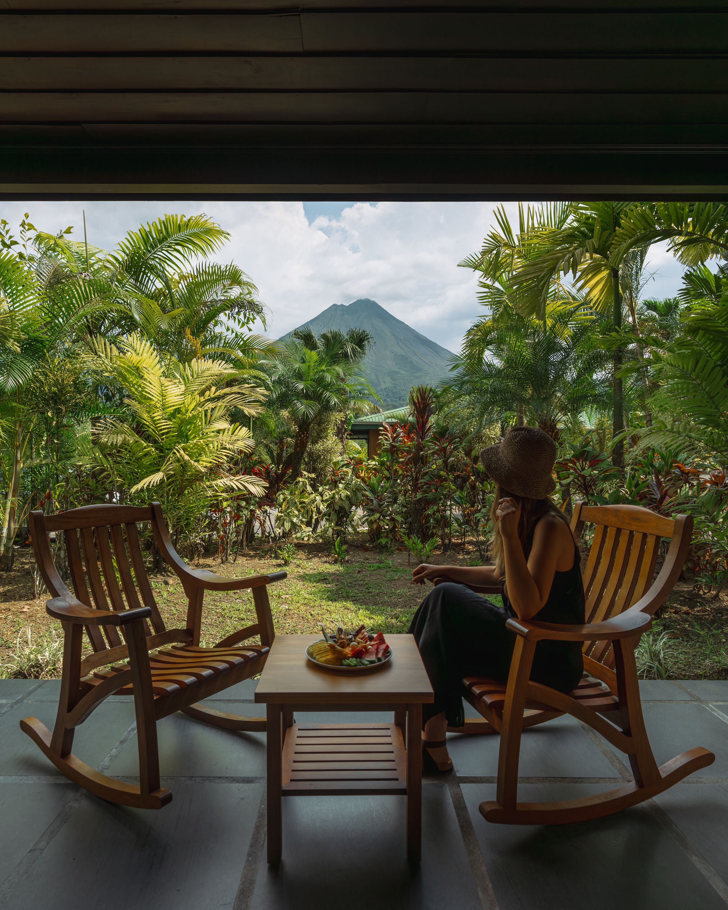 A person sitting on a wooden rocking chair facing a table with a plate of food, overlooking a lush garden with a mountain in the background.