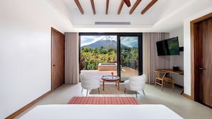 A modern hotel room with a large bed, two white chairs, a small table, a TV, and a balcony with a mountain view.
